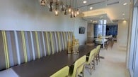 This thoughtfully designed single-floor space exudes a modern, upscale ambiance that blends comfort with contemporary style. The dining area serves as a central highlight, featuring a long, dark-topped communal table flanked by chartreuse upholstered chairs and a bold, striped high-back banquette in grey and yellow-green tones. Above, a dramatic cluster chandelier of mixed glass globe and elongated pendant lights creates a striking focal point, casting a warm, inviting glow throughout the space. The neutral grey walls and recessed lighting contribute to a clean, polished aesthetic. A flat-screen television is mounted on the wall, adding a functional element to the lounge-like setting. Glass partitions and walls extend the sightlines deeper into the space, revealing additional seating areas that suggest the layout is designed for both intimate gatherings and larger social settings. Natural light filters in through floor-to-ceiling windows visible in the background, enhancing the open, airy feel of the interior. The overall design palette — combining soft neutrals with pops of yellow-green and warm metallic accents from the decorative vessels on the table — gives the space a cohesive, sophisticated character well-suited for dining, socializing, or collaborative gatherings.