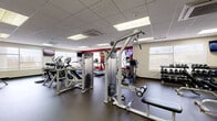 Located at 150 Blodgett Dr in Oneonta, NY, this well-appointed fitness center offers a generously sized, single-floor layout designed to accommodate a comprehensive workout experience. The main exercise floor features a dark rubber flooring surface ideal for high-traffic use, and is outfitted with a wide array of commercial-grade equipment. Cardio machines from Precor and Cybex — including treadmills, ellipticals, and stair climbers — line the perimeter, while multi-station cable machines, weight benches, and selectorized strength equipment occupy the central floor space. Multiple wall-mounted flat-screen televisions are positioned throughout the main area to keep users engaged during workouts. Large windows along the exterior walls invite natural light and provide outdoor views, contributing to an open and energizing atmosphere. A drop ceiling with recessed panel lighting ensures consistent, bright illumination throughout. A separate studio room features wood-look flooring, full-length mirrors, mounted televisions, a rack of colorful kettlebells, yoga mats, and free weights, making it ideal for stretching, yoga, or small-group functional training sessions. The overall design is clean, modern, and functional, providing a well-rounded fitness environment.