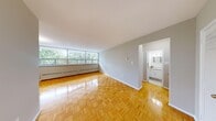 This 2-bedroom, 1-bathroom unit offers a well-organized layout that makes efficient use of its space. The living room is an immediate highlight, featuring warm parquet flooring in a distinctive basket-weave pattern that extends throughout the unit. A bank of large windows spans one wall, flooding the space with natural light and offering leafy treetop views that create a bright, airy ambiance. Baseboard heating runs beneath the windows, ensuring year-round comfort.

The kitchen is designed for functionality, outfitted with white cabinetry, light stone-finish countertops, a double-basin stainless steel sink, and white appliances including a refrigerator and range. White tile flooring and a classic tile backsplash complete the clean, bright aesthetic.

The primary bedroom is a well-proportioned retreat, benefiting from its own window with treetop views and the same polished parquet flooring. The second bedroom is equally bright and practical, with closet access and a connection to the hallway. The hallway provides a logical flow between rooms and includes thermostat controls.

The full bathroom features a tub-shower combination framed by clean white subway-style wall tiles, maintaining the unit's consistent, neutral palette throughout.