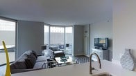 This single-floor space offers a thoughtfully designed layout that balances functionality and modern aesthetics. The entry hallway sets a stylish tone, featuring hardwood-style flooring, neutral grey walls, and a striking sunburst mirror that adds a decorative focal point. A large built-in storage unit lines one side of the corridor, while doorways on either end lead to the living and sleeping areas, creating a natural flow throughout the space.

The living area is bright and open, anchored by a curved arrangement of floor-to-ceiling windows that invite abundant natural light and offer expansive views of the surrounding urban landscape. The grey walls and smooth ceilings complement the modern furnishings, including a plush sectional sofa, accent armchair, and a reflective glass coffee table. A white media console with a mounted flat-screen television provides both storage and entertainment functionality.

The kitchen, visible from the living area, features a clean, contemporary design with a sleek countertop and a brushed gold faucet that adds a warm metallic accent. Overall, the space presents a cohesive, urban-modern ambiance with a neutral palette and refined finishes throughout.