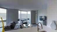 This single-floor space offers a thoughtfully designed layout that balances functionality and modern aesthetics. The entry hallway sets a stylish tone, featuring hardwood-style flooring, neutral grey walls, and a striking sunburst mirror that adds a decorative focal point. A large built-in storage unit lines one side of the corridor, while doorways on either end lead to the living and sleeping areas, creating a natural flow throughout the space.

The living area is bright and open, anchored by a curved arrangement of floor-to-ceiling windows that invite abundant natural light and offer expansive views of the surrounding urban landscape. The grey walls and smooth ceilings complement the modern furnishings, including a plush sectional sofa, accent armchair, and a reflective glass coffee table. A white media console with a mounted flat-screen television provides both storage and entertainment functionality.

The kitchen, visible from the living area, features a clean, contemporary design with a sleek countertop and a brushed gold faucet that adds a warm metallic accent. Overall, the space presents a cohesive, urban-modern ambiance with a neutral palette and refined finishes throughout.