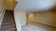 This two-story residence offers 3 bedrooms and 1.5 bathrooms across two well-organized floors. The main living area is a standout space, characterized by warm beige walls, light-toned tile flooring laid in a classic diagonal pattern, and a textured ceiling with recessed lighting that provides even, ambient illumination throughout. A notable architectural feature is the pair of arched alcoves built into the wall, each softly lit from within, adding a decorative focal point and a sense of depth to the room. White trim and baseboards provide clean contrast against the neutral walls, contributing to a cohesive, polished look. A carpeted staircase with a white balustrade leads to the upper floor, where the bedrooms and additional living spaces are located. The layout offers a practical flow between levels, with HVAC vents visible for climate control convenience. The overall ambiance of the main floor is open and inviting, with the arched details lending a subtle architectural character that distinguishes the space. The combination of recessed lighting, neutral finishes, and clean lines creates a versatile interior ready for a variety of furnishing styles.