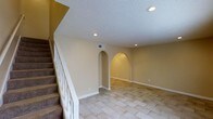 This two-story residence offers 3 bedrooms and 1.5 bathrooms across two well-organized floors. The main living area is a standout space, characterized by warm beige walls, light-toned tile flooring laid in a classic diagonal pattern, and a textured ceiling with recessed lighting that provides even, ambient illumination throughout. A notable architectural feature is the pair of arched alcoves built into the wall, each softly lit from within, adding a decorative focal point and a sense of depth to the room. White trim and baseboards provide clean contrast against the neutral walls, contributing to a cohesive, polished look. A carpeted staircase with a white balustrade leads to the upper floor, where the bedrooms and additional living spaces are located. The layout offers a practical flow between levels, with HVAC vents visible for climate control convenience. The overall ambiance of the main floor is open and inviting, with the arched details lending a subtle architectural character that distinguishes the space. The combination of recessed lighting, neutral finishes, and clean lines creates a versatile interior ready for a variety of furnishing styles.