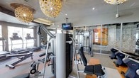This single-floor fitness center offers a comprehensive workout environment designed to accommodate a variety of exercise routines. The space is outfitted with an array of strength training equipment, including a multi-function cable machine, a functional trainer, and various resistance machines, alongside cardio equipment such as a stationary bike visible toward the perimeter. A ceiling-mounted DirecTV screen provides entertainment options for those exercising. The room features a combination of recessed lighting and distinctive decorative pendant lights with ornate, floral-inspired shades, lending a stylish, upscale ambiance uncommon in typical gym settings. Large glass panel windows and partitions along one wall allow natural light to filter into the space and provide visual openness. An accent wall featuring stacked stone detailing adds a warm, textural contrast to the otherwise modern interior. The neutral-toned flooring and light gray walls contribute to a clean, contemporary aesthetic, making this fitness center both functional and visually appealing.
