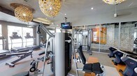 This single-floor fitness center offers a comprehensive workout environment designed to accommodate a variety of exercise routines. The space is outfitted with an array of strength training equipment, including a multi-function cable machine, a functional trainer, and various resistance machines, alongside cardio equipment such as a stationary bike visible toward the perimeter. A ceiling-mounted DirecTV screen provides entertainment options for those exercising. The room features a combination of recessed lighting and distinctive decorative pendant lights with ornate, floral-inspired shades, lending a stylish, upscale ambiance uncommon in typical gym settings. Large glass panel windows and partitions along one wall allow natural light to filter into the space and provide visual openness. An accent wall featuring stacked stone detailing adds a warm, textural contrast to the otherwise modern interior. The neutral-toned flooring and light gray walls contribute to a clean, contemporary aesthetic, making this fitness center both functional and visually appealing.