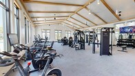 Located at 2900 Height St in Sarasota, FL, this fitness center is a standout facility defined by its striking architectural design and generous floor area. The space features a dramatic vaulted ceiling with exposed natural wood beams and white shiplap paneling, creating an open and airy atmosphere that sets it apart from a typical gym environment. Large black-framed windows line the walls on multiple sides, flooding the interior with natural light and offering pleasant outdoor views of the surrounding Florida landscape, including palm trees and the broader community.

The flooring throughout is durable, low-pile carpet tile in a neutral gray tone, providing comfort and traction across the entire workout area. The facility is comprehensively equipped to support a wide range of fitness goals. Cardio equipment includes Echelon stationary bikes, TRUE brand treadmills, ellipticals, and stair climbers, all arranged along the windowed walls to take advantage of the natural views. The strength training area features a robust selection of Instinct-brand selectorized machines, cable systems, free-weight benches, a dedicated dumbbell rack, and functional training rigs with accessories such as punching bags and suspension trainers. Full-length mirrors along one wall visually expand the space and support proper form during workouts. Ceiling fans and pendant lighting complete the comfortable, well-appointed interior.