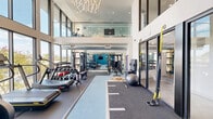 Located at 525 N 7th St in San Jose, CA, this well-appointed fitness center offers a thoughtfully designed exercise space with an impressive double-height ceiling that creates an open, airy atmosphere. A bold teal accent wall anchors the interior, adding a vibrant energy to the room, while large floor-to-ceiling windows along one side flood the space with abundant natural light and offer views of the surrounding area. A striking chandelier overhead adds a contemporary design touch rarely seen in fitness facilities.

The exercise floor is equipped for a wide variety of workout styles. Cardio equipment includes treadmills, a StairMaster, and a WaterRower, while the strength training zone features a multi-station cable machine, a full dumbbell rack ranging across multiple weights, kettlebells, and a flat bench. Functional training is well represented with TRX suspension trainers, battle ropes, resistance bands, and a stability ball. A central turf-style runner defines an open functional training lane ideal for dynamic movements.

The flooring transitions between rubberized exercise mats and low-pile carpet zones, catering to different training needs. A wall-mounted flat-screen TV provides entertainment. The glass-panel entry doors connect the gym seamlessly to adjacent building amenities, and an upper mezzanine overlooks the space, further enhancing the facility's sense of scale and modern design.