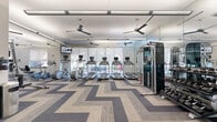 Located at 2950 Old Spanish Trail in Houston, TX, this fitness center offers a comprehensive, single-floor exercise facility designed to support a full range of workout routines. The space is generously sized, providing ample room for both cardio and strength training activities without feeling crowded.

The cardio zone is anchored by a row of treadmills equipped with individual touchscreen monitors, complemented by elliptical trainers and other aerobic machines. Large floor-to-ceiling windows line the perimeter, flooding the space with natural light and offering views of the outdoor surroundings. Solar shades allow for glare control while maintaining brightness throughout the room.

The strength training area features a multi-station cable machine, a dumbbell rack with a wide weight selection, and various resistance machines to target all major muscle groups. A dedicated section accommodates functional fitness equipment, including benches, stability balls, and additional free weights.

Full-length mirrors are strategically placed along the walls, enhancing the sense of openness and allowing users to monitor their form. Ceiling fans suspended from the white ceiling promote airflow and comfort during workouts. The floor is finished with a stylish chevron-patterned carpet tile in neutral tones of gray and beige, adding a modern, polished aesthetic. Linear LED lighting ensures consistent, bright illumination throughout the entire facility.