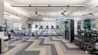 Located at 2950 Old Spanish Trail in Houston, TX, this fitness center offers a comprehensive, single-floor exercise facility designed to support a full range of workout routines. The space is generously sized, providing ample room for both cardio and strength training activities without feeling crowded.

The cardio zone is anchored by a row of treadmills equipped with individual touchscreen monitors, complemented by elliptical trainers and other aerobic machines. Large floor-to-ceiling windows line the perimeter, flooding the space with natural light and offering views of the outdoor surroundings. Solar shades allow for glare control while maintaining brightness throughout the room.

The strength training area features a multi-station cable machine, a dumbbell rack with a wide weight selection, and various resistance machines to target all major muscle groups. A dedicated section accommodates functional fitness equipment, including benches, stability balls, and additional free weights.

Full-length mirrors are strategically placed along the walls, enhancing the sense of openness and allowing users to monitor their form. Ceiling fans suspended from the white ceiling promote airflow and comfort during workouts. The floor is finished with a stylish chevron-patterned carpet tile in neutral tones of gray and beige, adding a modern, polished aesthetic. Linear LED lighting ensures consistent, bright illumination throughout the entire facility.