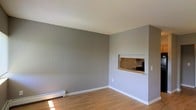 This single-floor residential space offers a clean, functional layout with a cohesive modern aesthetic. The living area is finished with warm light hardwood flooring that runs throughout, complemented by crisp white baseboards and smooth gray-painted walls that create a calm, neutral atmosphere. A textured popcorn ceiling adds a classic touch overhead, while a baseboard heater along the lower wall ensures practical comfort during cooler months.

A standout architectural detail is the built-in pass-through window that connects the living area to the kitchen, allowing for an open flow of light and communication between the two spaces. This recessed opening provides a glimpse into the kitchen area, which features cabinetry and countertop workspace visible through the cutout. The pass-through is framed neatly with a white surround, blending seamlessly with the overall décor.

The space is well-lit by natural light entering from the adjacent areas, enhancing the airy, open feel of the living room. The layout is straightforward and versatile, making it suitable for a variety of furnishing arrangements. Overall, the space presents a tidy, move-in-ready environment with practical features and a simple, understated design sensibility.
