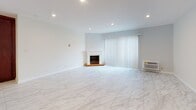 This single-floor residence offers a well-organized layout with clearly defined living areas, each finished with complementary materials and a clean, neutral palette throughout. The living room serves as a central gathering space, featuring large-format marble-look tile flooring, recessed lighting, and a classic fireplace with a white surround as a focal point. A sliding glass door dressed with vertical blinds allows natural light to fill the room while providing views of mature palm trees outside.

The kitchen is equipped with rich, dark mahogany-toned cabinetry extending floor to ceiling for generous storage capacity. White appliances, including an electric range, over-the-range microwave, and refrigerator, are set against light countertops and a tile backsplash. The layout flows efficiently, with a peninsula counter separating the kitchen from the adjacent dining or utility area.

The bedroom continues the warm aesthetic with rich hardwood flooring, recessed lighting, and its own sliding glass door with vertical blinds that frame views of lush outdoor greenery. A baseboard heating unit runs along the wall for added comfort.

The bathroom is finished with large-format stone-look tile on both the floor and shower surround, complemented by a white vanity with a quartz-style countertop and brushed nickel fixtures, creating a clean and polished finish.