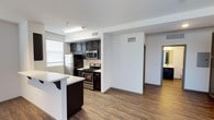 This single-floor space showcases a thoughtfully designed open-concept layout that seamlessly connects the kitchen and living areas. The kitchen is outfitted with dark espresso cabinetry that provides a striking contrast against the white countertops, including a breakfast bar with seating overhang — ideal for casual dining or entertaining. Stainless steel appliances, including a gas range, built-in microwave, and refrigerator, add a modern, functional touch to the space.

Wood-look plank flooring runs throughout the main living area, offering both durability and visual warmth. The neutral white walls and standard-height ceilings contribute to a clean, contemporary aesthetic. Overhead fluorescent lighting in the kitchen ensures well-lit meal preparation, while a window allows natural light to filter into the space.

A hallway visible from the main area leads to additional rooms, suggesting a practical, well-organized floor plan. Ceiling-mounted HVAC vents are positioned throughout, ensuring consistent climate control. Overall, the space presents a modern, move-in-ready environment with a cohesive design palette.