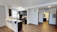 This single-floor space showcases a thoughtfully designed open-concept layout that seamlessly connects the kitchen and living areas. The kitchen is outfitted with dark espresso cabinetry that provides a striking contrast against the white countertops, including a breakfast bar with seating overhang — ideal for casual dining or entertaining. Stainless steel appliances, including a gas range, built-in microwave, and refrigerator, add a modern, functional touch to the space.

Wood-look plank flooring runs throughout the main living area, offering both durability and visual warmth. The neutral white walls and standard-height ceilings contribute to a clean, contemporary aesthetic. Overhead fluorescent lighting in the kitchen ensures well-lit meal preparation, while a window allows natural light to filter into the space.

A hallway visible from the main area leads to additional rooms, suggesting a practical, well-organized floor plan. Ceiling-mounted HVAC vents are positioned throughout, ensuring consistent climate control. Overall, the space presents a modern, move-in-ready environment with a cohesive design palette.