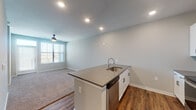 This thoughtfully designed single-floor unit offers a clean, contemporary aesthetic throughout. The open-concept layout connects the kitchen and living area seamlessly, creating a spacious and functional flow. The kitchen is a standout feature, equipped with white shaker-style cabinetry, quartz countertops, a decorative tiled backsplash, and a full suite of stainless steel appliances including a side-by-side refrigerator with water and ice dispenser, an electric range, an over-the-range microwave, and a dishwasher. A central island with a built-in sink adds both prep space and visual appeal. Rich wood-look flooring extends through the kitchen and main living areas, providing a warm contrast to the light gray walls and bright recessed lighting overhead. The living area is generously sized, enhanced by a ceiling fan and a large window fitted with blinds that invites natural light and outdoor views. The bedroom continues the soft, comfortable feel with plush carpeting, a ceiling fan, and a sizable window overlooking the surroundings. The bathroom is well-finished with a white vanity, quartz countertop, a bold-framed mirror, wall-mounted lighting, and additional upper cabinetry for storage. Wood-look flooring carries through the bathroom, maintaining a cohesive look throughout the unit.