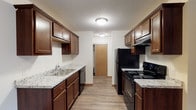 This single-floor space is defined by a thoughtfully designed galley-style kitchen that serves as its centerpiece. The kitchen features rich, dark wood cabinetry extending from floor to ceiling on both sides, providing generous storage throughout. Granite-look countertops in a light gray and white pattern run along both walls, offering ample prep space. The kitchen is equipped with black appliances, including an electric range with a built-in overhead exhaust hood and a full-sized refrigerator, as well as a stainless steel dishwasher seamlessly integrated beneath the counter. A double stainless steel sink is positioned along one wall, complemented by a standard faucet. The flooring throughout is a warm-toned laminate wood plank, adding a cohesive and durable finish to the space. Flush-mount ceiling light fixtures provide consistent, practical illumination, while a textured ceiling adds subtle visual character. At the far end of the kitchen, a natural wood-toned door leads to an adjacent area, suggesting a well-connected and functional layout. The overall ambiance is clean, practical, and welcoming, with a neutral color palette that balances the warmth of the wood cabinetry.