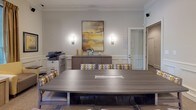 This single-floor business center is thoughtfully designed to accommodate a variety of professional needs within a cohesive, elegantly styled environment. The space is defined by consistent classical architectural detailing, including crown molding, wainscoting panels, and white columns that lend a refined, polished character throughout.

At the heart of the space is a generously sized conference room centered around a large, contemporary gray wood-grain table surrounded by upholstered chairs featuring a bold geometric pattern in gold and gray tones. A credenza along the back wall is flanked by wall sconces and accented by a large framed abstract artwork, while a multifunction printer/copier adds practical utility. The integrated cable management in the table surface supports seamless meetings and presentations.

A dedicated row of individual computer workstations lines one wall, separated by white columns and framed mirrors that enhance the sense of depth and light. Each station is equipped with a desktop monitor and ergonomic seating, offering private, focused work areas.

A comfortable lounge nook provides a more relaxed setting, furnished with cushioned armchairs in warm gold tones, a side table with a modern lamp, and large windows dressed with wooden blinds and drape panels that invite natural light and views of lush greenery outside. Soft neutral carpet, recessed lighting, and a warm, muted color palette tie the entire space together into a professional yet welcoming atmosphere.