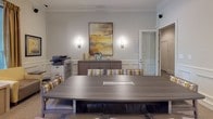 This single-floor business center is thoughtfully designed to accommodate a variety of professional needs within a cohesive, elegantly styled environment. The space is defined by consistent classical architectural detailing, including crown molding, wainscoting panels, and white columns that lend a refined, polished character throughout.

At the heart of the space is a generously sized conference room centered around a large, contemporary gray wood-grain table surrounded by upholstered chairs featuring a bold geometric pattern in gold and gray tones. A credenza along the back wall is flanked by wall sconces and accented by a large framed abstract artwork, while a multifunction printer/copier adds practical utility. The integrated cable management in the table surface supports seamless meetings and presentations.

A dedicated row of individual computer workstations lines one wall, separated by white columns and framed mirrors that enhance the sense of depth and light. Each station is equipped with a desktop monitor and ergonomic seating, offering private, focused work areas.

A comfortable lounge nook provides a more relaxed setting, furnished with cushioned armchairs in warm gold tones, a side table with a modern lamp, and large windows dressed with wooden blinds and drape panels that invite natural light and views of lush greenery outside. Soft neutral carpet, recessed lighting, and a warm, muted color palette tie the entire space together into a professional yet welcoming atmosphere.