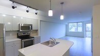 This single-floor space showcases a thoughtfully designed open-concept layout where the kitchen flows seamlessly into the living area. The kitchen is equipped with stainless steel appliances, including a range and over-the-range microwave, complemented by sleek white cabinetry offering ample storage. A curved kitchen island with a white countertop and stainless steel sink serves as a functional centerpiece, ideal for both meal preparation and casual dining. Two cylindrical pendant lights hang above the island, providing focused task lighting, while a track lighting system overhead adds further illumination to the kitchen workspace. The cabinetry features a clean, minimalist aesthetic in a light gray-white finish. The open living area beyond the kitchen benefits from natural light streaming through a large window, offering views of surrounding greenery. The hard-surface flooring throughout the space contributes to a cohesive, contemporary feel. The neutral color palette of soft whites and grays creates a bright, airy atmosphere, making the space feel open and inviting.