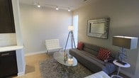 This well-appointed two-story space showcases a modern, contemporary design sensibility with a cohesive aesthetic carried throughout. The living area sets the tone with neutral-toned walls that create a clean, airy backdrop for the carefully curated furnishings. Sleek track lighting runs along the ceiling, providing both functional illumination and a modern architectural detail. Natural light filters in through a window fitted with horizontal blinds, brightening the space. The living room features a plush gray sofa accented with bold red geometric-patterned throw pillows, paired with a classic white tufted Barcelona-style chair and a polished oval glass coffee table resting on a textured area rug. A decorative mirror with a mosaic-style frame anchors one of the accent walls, adding depth and visual interest. Light-toned hardwood flooring runs throughout, lending warmth and continuity to the space. A tripod floor lamp adds a sculptural, designer touch. The overall ambiance is modern and inviting, balancing comfort with contemporary style across both floors, making it a versatile and thoughtfully designed space.