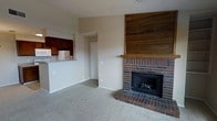 This single-story space offers a comfortable, open-concept layout that connects the living area and kitchen seamlessly. The living room serves as the focal point of the space, anchored by a classic brick fireplace with a raised hearth and a built-in wood mantel. Above the fireplace, a natural wood plank accent wall adds warmth and texture, creating a rustic yet inviting atmosphere. The living area is finished with neutral-toned carpet, contributing to a cozy and understated aesthetic throughout.

The kitchen is accessible from the living area and features a raised bar-style counter that functions as a natural divider between the two spaces while maintaining an open feel. Dark wood cabinetry lines the kitchen walls, providing ample storage, and standard appliances including a refrigerator and microwave are present. The cabinetry's rich tones complement the wood accents found in the living area, creating a cohesive design throughout. A doorway visible from the main living area provides access to additional rooms. Neutral wall tones and consistent flooring materials tie the entire space together in a clean, functional design.
