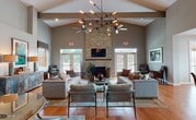 This single-story communal space is defined by its impressive architectural design, centered around a soaring vaulted ceiling with exposed beams that draw the eye upward. A striking floor-to-ceiling stacked-stone fireplace serves as the focal point of the main lounge area, topped with a wall-mounted television and flanked by two sets of French doors that flood the space with natural light and provide direct access to an outdoor patio. Overhead, a bold, multi-arm chandelier and ceiling fans complement the airy, open atmosphere.

The main lounge is generously proportioned, furnished with multiple tufted sofas, accent chairs, and layered area rugs atop warm hardwood flooring, creating a comfortable and inviting gathering environment. Additional seating nooks are positioned near the large windows, offering outdoor views and natural light throughout.

A separate dining area accommodates casual seating around a natural wood table with woven rattan chairs, positioned near its own set of French doors leading outside. The functional kitchen features white cabinetry, full appliances including a gas range and refrigerator, and tile flooring, providing practical support for events and gatherings. Together, these areas form a well-appointed, versatile community space designed for both relaxation and social interaction.