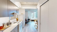 Located at 200 Folsom Street in San Francisco's Rincon Hill neighborhood, this efficient one-bedroom, one-bathroom unit offers a well-designed open floor plan across a single level. The living area and kitchen flow seamlessly together, with light wood-style flooring and warm wood cabinetry lining the kitchen, which is equipped with a full-size refrigerator, range, and ample upper and lower cabinet storage. A dedicated living zone accommodates a sofa and coffee table beneath a distinctive wood-paneled ceiling accent that adds warmth to the space. The bedroom area is highlighted by a striking corner window configuration that frames elevated city views of the San Francisco skyline, filling the room with abundant natural light. Exposed concrete columns provide an industrial architectural detail that contrasts with the otherwise clean, white interior walls. The bathroom features a wall-mounted vanity with cane-front cabinet storage, a large mirror with overhead lighting, and light wood-tone flooring consistent with the rest of the unit. The entrance includes a closet and additional storage space. The overall design balances compact, functional living with modern urban aesthetics.
