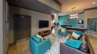 Located at 120 Plantation Creek Dr in Mooresville, NC, this single-floor community clubhouse delivers a well-appointed gathering space designed for both relaxation and productivity. The open layout flows seamlessly across multiple functional zones, all unified by warm hardwood-style flooring, bold teal accent walls, and rich brown trim that together create a cohesive, contemporary aesthetic.

The central game and lounge area serves as the heart of the space, anchored by a full-size pool table beneath a striking multi-bulb chandelier. Teal upholstered seating, patterned area rugs, and a wall-mounted flat-screen TV furnish the lounge zone, encouraging casual gathering. A dining-height table with dark chairs provides additional seating for group use.

A standout architectural feature is a floor-to-ceiling stacked stone column that anchors the business center area, where workstations with desktop computers are arranged for resident use. Pendant lighting illuminates the workspace, which is framed by a textured accent wall adding depth and visual interest.

A cozy nook with a round bistro table, leather chairs, decorative mirrors, and woven wall art creates an intimate setting for smaller conversations. Throughout, recessed lighting and carefully chosen décor contribute to a polished, inviting ambiance.