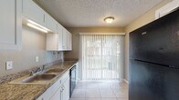 This single-floor residence offers a practical and well-organized layout with light-toned tile flooring running consistently throughout the space. The kitchen is a functional highlight, featuring white cabinetry, granite-style countertops, a double basin stainless steel sink, and black appliances including a dishwasher and refrigerator. A large sliding glass door with vertical blinds fills the kitchen with natural light while providing outdoor views. The textured ceilings are present throughout the home, lending a consistent architectural character to each room. The living and dining areas are open and versatile, with neutral gray-toned walls, exposed structural beams, and ample wall space suited to a variety of arrangements. Each bedroom is equipped with a ceiling fan and light fixture, and features windows that invite natural light. At least one bedroom includes a walk-in closet, offering generous storage. The bathroom is cleanly appointed with a full tub-shower combo, granite-style vanity countertop, white cabinetry, and tile flooring that mirrors the rest of the home. Overall, the space presents a clean, neutral palette that provides a functional and adaptable living environment.