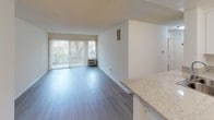 This single-floor residence offers a clean, modern aesthetic defined by light-toned wood-style flooring that flows seamlessly throughout the entire unit. The open-concept layout connects the living area to the kitchen, creating a cohesive and functional living environment. The living room is generously proportioned, bathed in natural light through large sliding glass doors dressed with vertical blinds that open to an outdoor view, lending the space an airy, inviting quality. A wall-unit air conditioner ensures year-round comfort.

The kitchen is a standout feature, outfitted with crisp white shaker-style cabinetry extending to the ceiling for ample storage, light granite-style countertops, and a peninsula with a double stainless steel sink — ideal for both cooking and casual dining. Stainless steel appliances, including a gas range and a full-size refrigerator, complete the well-appointed culinary space.

A separate bedroom offers a tranquil retreat with its own vertical-blind window framing a leafy outdoor view, while a textured ceiling adds subtle character. The entry hallway is clean and functional, illuminated by a ceiling-mount fixture and providing easy access to all areas of the unit. Overall, the space presents a fresh, well-maintained interior with a neutral palette ready to complement any style.