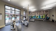 This well-equipped fitness center draws immediate attention with a bold, large-scale Nashville skyline mural spanning an entire accent wall, depicting the city's iconic silhouette in shades of blue, green, and gold against a distressed typographic backdrop. The space is generously sized, providing ample room for a comprehensive range of exercise equipment without feeling crowded. Durable rubber flooring runs throughout, offering both safety and practicality underfoot.

The cardio zone features multiple treadmills, elliptical trainers, and stationary bikes, several of which are equipped with individual display consoles. Large floor-to-ceiling windows with black-framed grid detailing line one side of the room, inviting natural light and providing views of the outdoor pool area and surrounding property. The bright, open feel is further enhanced by overhead fluorescent lighting mounted to a smooth, neutral-toned ceiling.

Strength training is well represented with a dedicated weight area that includes a dumbbell rack with a full range of free weights, a flat utility bench, and multiple selectorized weight machines. A wall-mounted flat-screen television is positioned for visibility across the workout floor. Wall mirrors provide visual expansion and functional utility. The overall aesthetic balances Nashville local character with a clean, modern, and functional design.