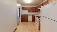 This single-floor residential unit offers a practical and well-organized layout designed for comfortable everyday living. The kitchen serves as a functional hub, featuring warm oak cabinetry with ample upper and lower storage, light-toned countertops, tile flooring, and white appliances including an electric range, dishwasher, and refrigerator. A double-basin sink and a range hood complete the space, which opens toward the rest of the unit.

The living and bedroom areas are finished with soft, neutral wall-to-wall carpeting and white walls accented by wood baseboards. Large windows dressed with vertical blinds allow generous natural light to fill the rooms while offering views of outdoor greenery. A wall-mounted air conditioning unit and baseboard heating provide year-round climate control. One room features an elegant chandelier with a brass finish, adding a decorative touch to the space.

The bathroom is clean and functional, equipped with tile flooring, a wood-toned vanity, a toilet, and a tub/shower combination fitted with safety grab bars. The walk-in closet provides notable storage capacity, featuring dual hanging rods and shelving on both sides, making it a practical asset to the unit. Textured ceilings are present throughout, contributing to the overall classic residential character of the space.
