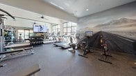 This modern fitness center is designed to support a comprehensive workout experience within a thoughtfully organized, single-floor layout. The space is divided into distinct functional zones, each catering to different aspects of physical training. The cardio area is highlighted by a row of treadmills, ellipticals, and stationary bikes — including interactive cycling machines — all positioned in front of expansive floor-to-ceiling windows that offer abundant natural light and direct views of the outdoor pool and surrounding courtyard. A dramatic photographic landscape mural of a desert highway and mountain range serves as a striking visual backdrop, adding an immersive quality to the environment. The strength training zone is well-appointed with a multi-function cable machine, an adjustable weight bench, a full rack of dumbbells, medicine balls, and a large wall-mounted mirror paired with a flat-screen TV. Rubberized flooring throughout provides both safety and durability. The space also features high ceilings in the strength area, enhancing the sense of openness. Glass-paned interior doors and direct access to the outdoor courtyard contribute to a bright, airy ambiance. Ceiling fans and recessed lighting complete the clean, contemporary aesthetic of this fully equipped fitness facility.