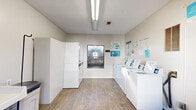 This shared laundry facility at 2845 Zelda Rd in Montgomery, Alabama, offers a clean, functional, and efficiently organized space designed to meet residents' laundry needs. The single-room layout is finished with light wood-look vinyl flooring and neutral-toned walls, creating a bright and welcoming environment. Overhead fluorescent lighting ensures the space is well-lit throughout.

Along one wall, a row of modern top-loading washing machines, managed through a CSC payment system, lines the room. Stacked dryer units are positioned nearby, maximizing the use of vertical space. A large, white folding table supported by hairpin legs provides a dedicated surface for sorting and folding laundry, while a utility sink mounted to the wall offers added convenience for hand-washing or stain treatment. A waste bin and iron hook are also present for practical use.

A window along one wall allows natural light to filter into the space, adding to its clean and airy feel. Informational posters and laundry tips are displayed on the walls, offering guidance on machine usage and best practices. The room is accessed through a solid entry door with a glass panel. The overall layout is straightforward and user-friendly, making this laundry facility a practical and well-maintained amenity.