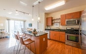 This single-floor residence offers a thoughtfully designed open-concept layout that seamlessly connects the kitchen, dining, and living areas. The kitchen is a standout feature, equipped with stainless steel appliances including a gas range, built-in microwave, and dishwasher, all complemented by rich dark granite countertops, warm medium-toned wood cabinetry, and hardwood flooring. A central island with a black granite surface and bar seating provides an ideal space for casual dining, illuminated by stylish pendant lighting overhead. The living area is bathed in natural light, courtesy of expansive floor-to-ceiling sliding glass doors that open to outdoor views of the surrounding landscape, creating a bright and airy atmosphere. The space includes multiple bedrooms, each featuring plush carpeting, generous natural light from well-positioned windows with blinds and curtains, and a calm, neutral color palette that promotes a restful environment. The bathroom is finished with warm wood vanity cabinetry, a white countertop, and a large mirror accented by elegant overhead lighting. Throughout the residence, cream-toned walls, consistent finishes, and a cohesive design language contribute to a comfortable and functional living environment.