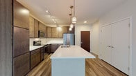 This single-floor residence showcases a thoughtfully designed open layout, with the kitchen serving as a clear centerpiece. The kitchen is outfitted with rich, dark wood cabinetry extending to the ceiling, offering generous storage capacity. White quartz countertops provide a crisp contrast, while a classic subway tile backsplash adds texture to the space. A substantial kitchen island with an undermount sink and pendant lighting above anchors the room, creating both a functional workspace and a natural gathering point. Stainless steel appliances, including a refrigerator, range, and microwave, complete the culinary setup. Wood-look plank flooring runs throughout the kitchen and living areas, adding warmth and continuity.

The bedrooms are finished with neutral-toned carpet, contributing to a quiet and comfortable atmosphere. Large black-framed windows allow natural light to fill these rooms while providing views of the surrounding exterior. A ceiling fan in one bedroom enhances comfort year-round.

The bathroom features light large-format tile flooring, a sliding glass shower enclosure, and a dark wood vanity with a white countertop, maintaining the same modern aesthetic found throughout the residence. Overall, the space presents a clean, contemporary design with consistent finishes.