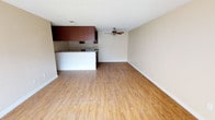 This single-floor space features an open-concept design that seamlessly integrates the living area with the kitchen. Light-toned hardwood-style flooring extends throughout, lending warmth and visual continuity to the space. The living area is generously proportioned, offering ample room for flexible furniture arrangements, and is equipped with a ceiling fan that contributes to both comfort and air circulation.

The kitchen is defined by a white peninsula counter that serves as a natural divider between the cooking and living zones. Rich, dark reddish-brown cabinetry is mounted overhead, providing a bold contrast against the neutral beige walls. Stainless steel appliances, including what appears to be a range and microwave, are integrated into the kitchen space, adding a functional and modern touch.

The ceilings feature a textured popcorn finish, and white baseboards frame the perimeter of the room cleanly. The overall palette — neutral walls, warm flooring, and dark cabinetry accents — creates a balanced and practical interior. The open layout maximizes the sense of space, making the unit feel bright and connected throughout.