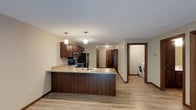 This single-floor residence offers a well-designed open-concept layout centered around a functional kitchen. The kitchen is outfitted with rich, dark wood cabinetry that provides ample storage, complemented by granite countertop surfaces that lend a polished, durable finish. A peninsula-style counter extends outward, creating an ideal space for casual dining or meal prep while maintaining visual connectivity to the surrounding living area.

Stainless steel appliances, including a refrigerator, microwave, and range, are seamlessly integrated into the cabinetry. Pendant lighting fixtures hang above the kitchen area, providing focused task lighting and adding a decorative touch to the space.

The flooring throughout is a light-toned wood-look material that adds warmth and continuity across the open layout. Neutral-toned walls keep the interior feeling bright and versatile. A hallway leads toward the unit's entry door and additional rooms beyond.

A notable feature of this space is the in-unit laundry, visible through an open doorway, offering added convenience. The overall design balances practicality with a clean, modern aesthetic.