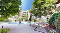 Located at 153 Valley St in South Orange, NJ, this thoughtfully designed outdoor amenity space serves as a serene courtyard retreat surrounded by a multi-story residential building. At its centerpiece sits a kidney-shaped swimming pool with accessible lift equipment, flanked by a generous sun deck outfitted with lounge chairs and umbrellas for relaxing under open skies. The pool area is framed by lush, mature trees that provide natural shade and a sense of seclusion.

Winding concrete pathways connect the various zones of the courtyard, guiding residents through carefully maintained garden beds filled with ferns, flowering plants, and ornamental shrubs. A raised wood deck with navy blue metal railings offers an inviting area for outdoor seating, surrounded by verdant greenery that creates a tranquil, park-like atmosphere. Adjacent to the deck, a neatly trimmed lawn area provides additional open green space, ideal for leisure and recreation.

Outdoor seating clusters are positioned throughout the courtyard, including areas near a pergola and grill stations, making the space well-suited for both casual gatherings and quiet relaxation. The multi-story building facade, combining warm yellow tones with contrasting dark brick and private balconies, encloses the courtyard and contributes to its intimate, community-focused ambiance.