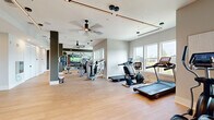 This well-appointed fitness center is organized across a single level, offering a comprehensive range of equipment and training zones designed to accommodate diverse workout routines. The space is finished with light hardwood-style flooring throughout, contributing to a clean, contemporary aesthetic that is both functional and visually inviting.

The cardio zone is positioned along a window-lined wall, where Spirit-brand treadmills, stationary bikes, and elliptical trainers benefit from generous natural light and outdoor views. Ceiling fans are installed throughout, ensuring comfortable airflow during workouts. Sleek linear pendant lighting runs along the ceiling, complementing the modern design.

The strength training section features a comprehensive selection of Hoist selectorized machines and a full dumbbell rack displayed in front of a large flat-screen television and mirrored wall, creating an open, energizing environment.

A dedicated functional training room offers additional versatility, equipped with a heavy bag, battle ropes, medicine balls, resistance equipment, and open floor space for dynamic movement. This area features a motivational wall graphic and a dark accent wall, distinguishing it visually from the main floor. Structural columns with gray accents punctuate the space, adding architectural interest throughout.