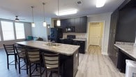 This single-floor unit is designed with a functional, modern aesthetic that suits shared living arrangements. The open-concept kitchen serves as a central hub, outfitted with dark espresso cabinetry, granite-style countertops, and black appliances including a range, microwave, and refrigerator. A spacious kitchen island with an integrated double sink and bar seating provides additional workspace and a casual dining option. Pendant lighting above the island and flush-mount fixtures throughout keep the space well-lit. Light wood-look flooring runs through the kitchen, hallways, and common areas, lending a clean, cohesive look.

Adjacent to the kitchen, a dedicated laundry area houses a full-size washer and dryer, adding everyday convenience. Extended countertop space along one kitchen wall offers additional storage and prep area.

The living area is bright and welcoming, with large windows that invite natural light and a relaxed atmosphere for gathering.

Multiple bedrooms provide private retreats, each furnished with a bed, desk, and chair — practical setups suited for study and rest. Some rooms feature carpet flooring, while others continue the wood-look finish. Ceiling fans and windows with blinds add comfort.

The bathroom features a dual-sink vanity with dark cabinetry, granite-style countertops, and a shower-tub combination, maintaining the unit's consistent modern design language throughout.