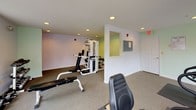 This fitness center offers a comprehensive workout environment designed to accommodate a variety of exercise routines. The space is divided into distinct functional zones, making it easy to transition between cardio training and strength work. The cardio area is positioned near a bank of large windows that allow generous natural light to fill the room, creating an open and energizing atmosphere. Equipment in this section includes a treadmill, stationary bike, and elliptical trainer, each placed on protective floor mats atop wall-to-wall carpeting. The strength training area features a multi-function cable and weight machine, a flat workout bench, and a rack of dumbbells in graduated sizes, providing options for both compound and isolated exercises. The walls are painted in soft lavender and sage green tones, lending the space a calm, welcoming ambiance. Recessed ceiling lighting supplements the natural light throughout, ensuring the room remains well-lit at all times. An emergency exit door and clearly marked safety signage reflect a commitment to user safety. The single-floor layout keeps everything accessible and easy to navigate, making this fitness center a practical and inviting space for daily exercise.