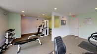 This fitness center offers a comprehensive workout environment designed to accommodate a variety of exercise routines. The space is divided into distinct functional zones, making it easy to transition between cardio training and strength work. The cardio area is positioned near a bank of large windows that allow generous natural light to fill the room, creating an open and energizing atmosphere. Equipment in this section includes a treadmill, stationary bike, and elliptical trainer, each placed on protective floor mats atop wall-to-wall carpeting. The strength training area features a multi-function cable and weight machine, a flat workout bench, and a rack of dumbbells in graduated sizes, providing options for both compound and isolated exercises. The walls are painted in soft lavender and sage green tones, lending the space a calm, welcoming ambiance. Recessed ceiling lighting supplements the natural light throughout, ensuring the room remains well-lit at all times. An emergency exit door and clearly marked safety signage reflect a commitment to user safety. The single-floor layout keeps everything accessible and easy to navigate, making this fitness center a practical and inviting space for daily exercise.