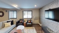 This single-floor space is designed with an open-concept layout that seamlessly connects a fully equipped kitchen, a comfortable lounge area, and a game room. The kitchen is a standout feature, showcasing rich dark cabinetry paired with light granite countertops, a stainless steel range, and a mosaic tile backsplash that extends behind a wall-mounted flat-screen television. A large granite-topped island provides additional workspace and casual seating. Warm, earthy tones on the kitchen walls complement the wood-look flooring that flows consistently throughout the entire space.

The lounge area is thoughtfully furnished with dark leather armchairs accented by rust-colored pillows, a light sectional sofa, and a plush area rug. A textured, patterned accent wall serves as a focal point for another wall-mounted flat-screen television. Windows along one wall allow natural light to filter in. The game area, visible in the floor plan, includes a billiard table, adding a recreational dimension to the space. Recessed lighting throughout ensures even illumination, while the cohesive design elements — including consistent flooring, neutral walls, and modern furnishings — create a unified and inviting atmosphere.
