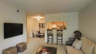 This single-floor residence features an open-concept design that seamlessly connects the living, dining, and kitchen areas into one cohesive space. The living room is carpeted and comfortably proportioned, with a wall-mounted flat-screen television and a low-profile coffee table serving as central focal points. A plush sofa and side table complete the seating arrangement, creating a relaxed and functional atmosphere.

The kitchen is positioned behind a half-wall pass-through, which doubles as a breakfast bar fitted with two cross-back bar stools. Warm wood-toned upper cabinetry lines the kitchen walls, and a range with an overhead hood is visible through the opening. The pass-through design enhances the open feel while maintaining a defined kitchen zone.

Beyond the kitchen pass-through, the dining area is clearly visible and features a dark-finished dining table surrounded by upholstered chairs. A ceiling fan with a light fixture illuminates this space, providing both airflow and ambient lighting. The textured popcorn ceiling spans the entire unit, and neutral wall tones throughout contribute to a cohesive, understated aesthetic. Soft carpeting runs through the living and dining areas, adding warmth underfoot.