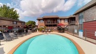 This community amenity space combines outdoor relaxation with indoor comfort, offering residents a well-rounded gathering destination. At its heart is a generously sized, kidney-shaped swimming pool with clear turquoise water, surrounded by a spacious concrete deck furnished with lounge chairs and shaded seating areas beneath a red umbrella. The pool area is enclosed by a wrought-iron fence and set against the backdrop of a classic two-story brick residential building.

The adjoining clubhouse interior is thoughtfully designed and decorated with a cohesive palette of sage green walls, warm hardwood flooring, and crown molding throughout. The main lounge area features plush leather seating arranged around a large flat-screen television, ceiling fans overhead, and recessed lighting that creates a welcoming ambiance. Framed abstract artwork adds a modern touch to the walls.

A dedicated dining area provides multiple round tables with cushioned chairs, ideal for casual gatherings. A short staircase leads to an upper lounge space with additional seating and a workstation. The kitchenette is a standout feature, boasting dark espresso cabinetry, granite countertops, a striking mosaic tile backsplash, pendant lighting, and stainless steel appliances including a microwave, toaster oven, and water dispenser.