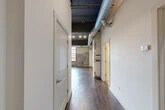 Located at 220 Herald Pl in Syracuse, NY, this industrial-style loft blends historic character with modern finishes across a single, well-appointed floor. The open living area immediately impresses with its soaring dark-painted ceilings, exposed silver ductwork, and rich hardwood flooring that runs throughout the main living spaces. Original brick walls add raw, authentic texture, while oversized factory-style windows framed in deep red bathe the interior in abundant natural light and offer views of the surrounding cityscape.

The kitchen is a standout, featuring rich mahogany cabinetry, granite countertops, stainless steel appliances, and a breakfast bar with pendant lighting — ideal for both everyday living and entertaining. The spacious primary bedroom continues the industrial aesthetic with exposed brick accents, large multi-pane windows, and plush carpeting for comfort.

Two full bathrooms provide practical functionality: one offers a walk-in glass-enclosed shower with tile surround and dark vanity cabinetry, while the second features tile flooring, a granite-topped vanity, and a glass-paneled shower. Additional spaces include a hallway, two closets, a utility room, and an unclassified flex room. This loft offers a compelling urban living experience rooted in architectural heritage.