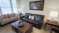 This single-floor space offers a warm and inviting atmosphere with a thoughtfully designed living area at its heart. The sitting room is anchored by a rich, dark leather sofa accented with blue and patterned throw pillows, complemented by a matching upholstered accent chair with a decorative print. A sturdy wooden coffee table with lower shelving serves as a functional centerpiece, while a small round side table holds a classic table lamp that adds a soft, ambient glow to the room. The walls are finished in a soft, neutral tone that enhances the sense of openness and calm. Natural light filters in through a window dressed with white blinds, brightening the space and adding an airy quality. A framed landscape artwork depicting a wooden bridge surrounded by autumn foliage adds a scenic focal point above the sofa, tying together the earthy, nature-inspired color palette throughout the room. The carpeted flooring adds to the overall coziness of the space, making it ideal for relaxed, everyday living. The overall design strikes a balance between comfort and understated elegance, making this a welcoming and functional living environment.