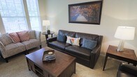 This single-floor space offers a warm and inviting atmosphere with a thoughtfully designed living area at its heart. The sitting room is anchored by a rich, dark leather sofa accented with blue and patterned throw pillows, complemented by a matching upholstered accent chair with a decorative print. A sturdy wooden coffee table with lower shelving serves as a functional centerpiece, while a small round side table holds a classic table lamp that adds a soft, ambient glow to the room. The walls are finished in a soft, neutral tone that enhances the sense of openness and calm. Natural light filters in through a window dressed with white blinds, brightening the space and adding an airy quality. A framed landscape artwork depicting a wooden bridge surrounded by autumn foliage adds a scenic focal point above the sofa, tying together the earthy, nature-inspired color palette throughout the room. The carpeted flooring adds to the overall coziness of the space, making it ideal for relaxed, everyday living. The overall design strikes a balance between comfort and understated elegance, making this a welcoming and functional living environment.