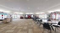 Located in Escondido, CA, this generously sized single-story rec room offers a welcoming and versatile community gathering space. The open floor plan encompasses a large main hall with a combination of smooth hardwood flooring and patterned carpet tiles, creating distinct zones for varied activities. A built-in service counter near the entrance provides a convenient focal point for events and gatherings, while a wall-mounted television and comfortable seating arrangements — including sofas, accent chairs, and small dining tables with chairs — make the space adaptable for both social and recreational use. A upright piano and a professional audio and lighting setup reflect the room's suitability for live performances and entertainment. Large glass double doors along one wall flood the interior with natural light and offer views of the outdoor surroundings, enhancing the overall brightness and openness of the space. Recessed lighting throughout the smooth white ceilings ensures consistent illumination across all areas. The rec room also features interior doors leading to additional utility and support spaces. With its expansive footprint, flexible layout, and well-appointed amenities, this rec room is well-equipped to host a wide range of community events, celebrations, and everyday social activities.