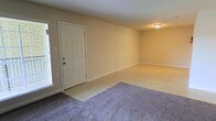 This single-story space presents a clean, functional layout with a welcoming entry area. The main living space is defined by a combination of light-toned ceramic tile flooring near the entryway and plush carpeting that extends through the primary living area, offering both practicality and comfort underfoot. The walls are finished in a warm beige tone, creating a neutral backdrop suitable for a variety of décor styles. A six-panel white entry door provides access to the space and is flanked by a window that introduces natural light, brightening the entry zone. White baseboards run along the perimeter of the room, adding a crisp contrast to the wall color. Overhead, a flush-mount ceiling light fixture provides ambient illumination. The open layout of the main room offers flexibility in furniture arrangement and use of space. Overall, the interior maintains a straightforward, well-maintained aesthetic with a light and airy feel, making it a versatile and functional single-floor space.